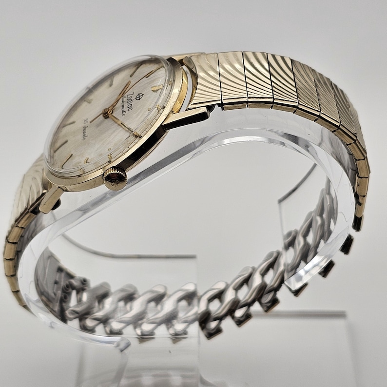 May include: A vintage Zodiac automatic wristwatch with a gold-toned case and a silver-colored dial. The watch features gold hands and hour markers, and a gold-toned expanding band. The watch is displayed on a clear stand.