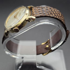 K&ouml;nnte beinhalten: Eine goldfarbene Armbanduhr mit cremefarbenem Zifferblatt und goldenen Zeigern. Die Uhr hat ein braunes Lederarmband mit einem strukturierten, schlangenartigen Muster auf dem oberen Teil. Die Uhr wird auf einem transparenten, gebogenen St&auml;nder pr&auml;sentiert.
