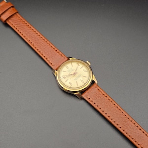 May include: A vintage gold-toned wristwatch with a light-colored face and brown leather band. The watch face displays numbers and hands, and the band is stitched. The watch is angled across a dark surface.