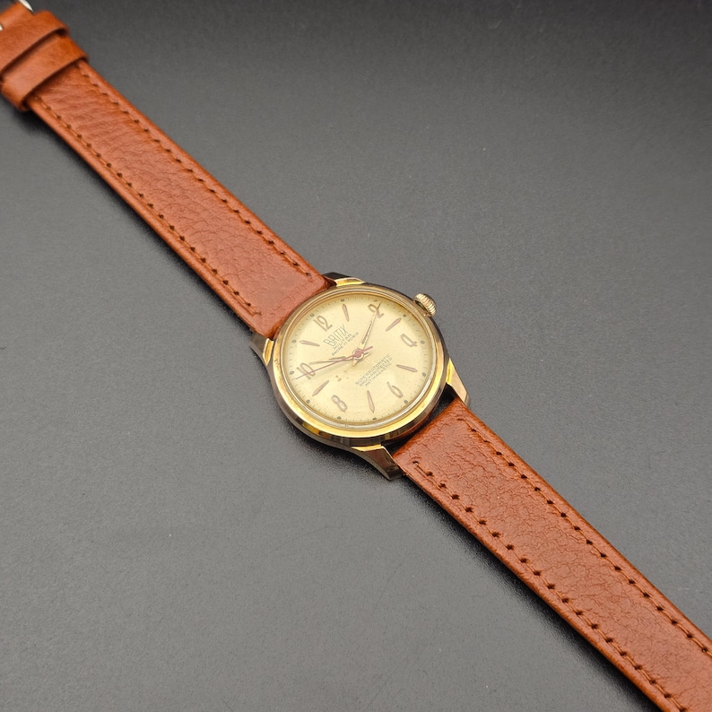 May include: A vintage gold-toned wristwatch with a light-colored face and brown leather band. The watch face displays numbers and hands, and the band is stitched. The watch is angled across a dark surface.