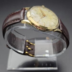 Pode incluir: Um rel&oacute;gio Zodiac dourado com mostrador claro e ponteiros dourados. O rel&oacute;gio tem uma pulseira de couro castanho com fivela dourada. O rel&oacute;gio &eacute; exibido em um suporte transparente e curvo.