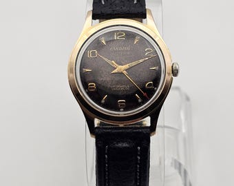May include: A vintage Candino wristwatch. It has a gold-tone case and a black leather strap. The watch face is dark with gold hands and numerals. The watch reads "Candino" and "17 Jewels Incabloc".