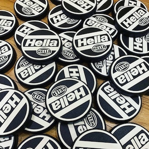 May include: A close-up of a pile of black and white round car emblems with the word "Hella" printed on them.