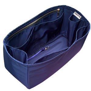 May include: A navy blue handbag organizer with multiple compartments and a zippered top. The interior is a lighter shade of blue, with a purple strap and gold-colored hardware. The brand name "PZEOTYN" is visible on a white tag.
