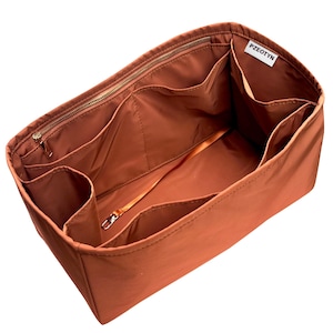 May include: A rust-colored handbag organizer with multiple compartments and a zippered top. The interior features various pockets and a strap with a metal clasp. The brand name "PZEOTYN" is visible on a small white tag.