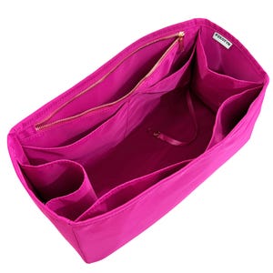 May include: A bright pink bag organizer with multiple compartments and a zippered closure. The interior features various pockets and a small strap with a clasp. The organizer is designed to fit inside a larger bag, providing organization and easy access to items.