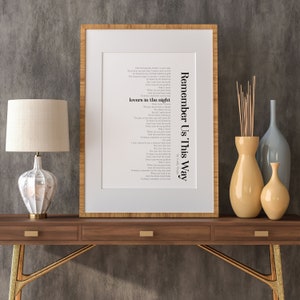 May include: A framed print of the song lyrics for "Remember Us This Way" by Lady Gaga. The lyrics are printed in black text on a white background. The frame is a light wood color. The print is sitting on a wooden table with two drawers. There are two yellow vases and one blue vase on the table.