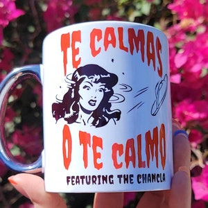 以下が含まれることがあります： 白いセラミック製のマグカップに、漫画の女性と「Te Calmas O Te Calmo Featuring The Chancla」という赤い文字と黒い文字が書かれています。