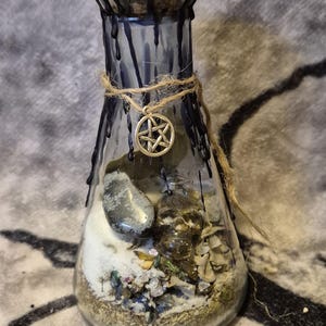 May include: A clear glass potion bottle with a black wax-sealed top and a gemstone. The bottle is adorned with a silver pentacle charm and twine. Inside are layers of sand, stones, and herbs, creating a mystical aesthetic.