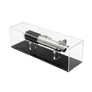 Peut inclure: Réplique de sabre laser argent et noir présentée dans un boîtier en acrylique transparent avec une base noire. Le sabre laser est détaillé avec divers composants métalliques et un bouton rouge. Le boîtier est rectangulaire.
