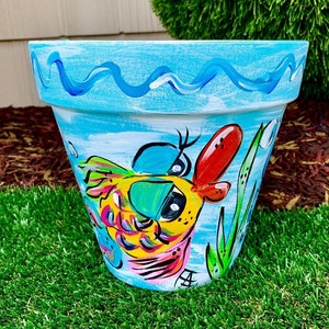 May include: Hand-painted ceramic flower pot with a whimsical fish design. The pot features a light blue background with a wavy border and a colorful fish illustration. The fish is yellow, pink, and blue, with red accents. Green grass and a brown mulch background.