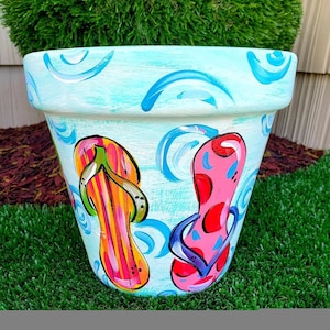 May include: A blue ceramic flower pot with a painted design of two colorful flip flops. The pot has a white background with blue swirls and the flip flops are pink, yellow, green, and purple.