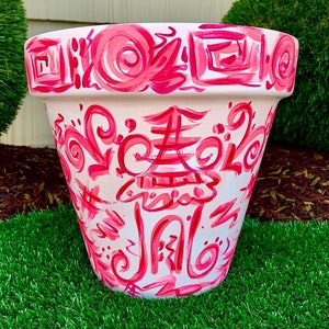 May include: A hand-painted, white ceramic flower pot with a pink and red design. The pot features a repeating pattern of swirls, geometric shapes, and a central pagoda-like illustration. The design is applied with visible brushstrokes, giving it a handcrafted look.