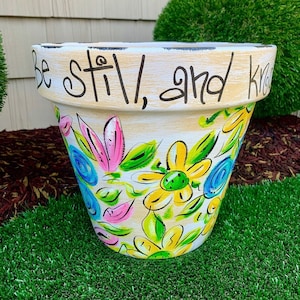 May include: A white ceramic flower pot with a hand-painted floral design in shades of pink, yellow, blue, and green. The pot has a black rim and the words "Be still, and know" written around the top.