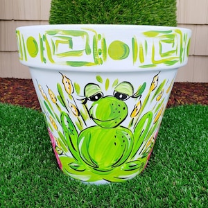 May include: White ceramic flower pot with a green frog painted on the front. The frog has eyelashes and is surrounded by yellow reeds. The pot has a green and white geometric design around the top rim.