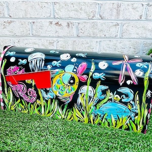 May include: Black mailbox with a hand-painted underwater scene. The design features colorful fish, seahorses, jellyfish, and a crab. The mailbox has a red flag and is trimmed with pink and white stripes.