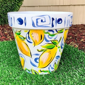 May include: White ceramic planter with a blue and white geometric design and hand-painted yellow lemons. The planter is sitting on a green artificial grass surface.