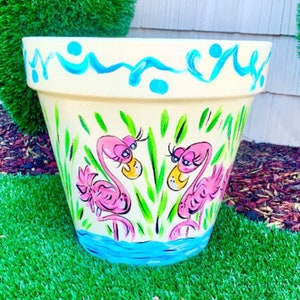 May include: A hand-painted terracotta planter with a light yellow background and a blue wavy border. Two pink flamingos are painted on the planter, with green leaves and a blue water scene at the bottom.