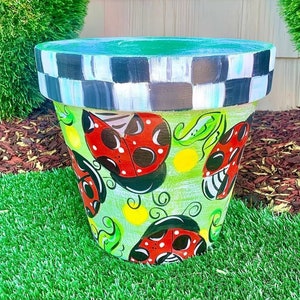 May include: A green and white painted terracotta planter with a checkered rim. The planter is decorated with a whimsical design of ladybugs, leaves, and yellow dots.