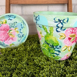 May include: Hand-painted ceramic flower pot and saucer set. The pot features a green bunny design with pink flowers and blue bows. The saucer has a matching floral pattern. The set is on a bed of green foliage.