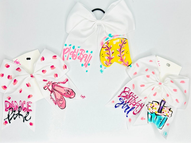 May include: Four white hair bows with pink polka dots. The bows are decorated with painted designs. One bow has a softball, one has a cupcake, one has ballet shoes, and one has the words 'Dance Like' written on it.