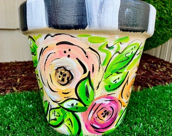 May include: A hand-painted flower pot with a cream base, adorned with pink, yellow, and orange roses and green leaves. The rim features black and white stripes. The pot is sitting on green grass.