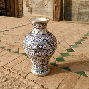 May include: A decorative vase with a silver rim and base, featuring intricate blue and white geometric patterns. The vase has a bulbous shape and is adorned with silver accents. It is placed on a brick surface.