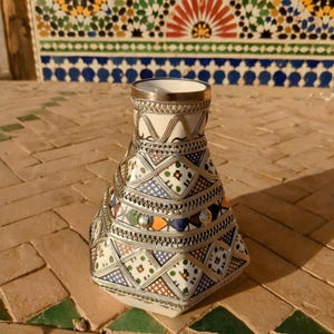 Może przedstawiać: Ozdobny ceramiczny wazon z białą podstawą i skomplikowanymi geometrycznymi wzorami w kolorze niebieskim, zielonym, pomarańczowym i brązowym. Wazon zdobiony jest srebrnymi metalowymi akcentami i srebrną obwódką. Wazon ma około 15 cm wysokości.