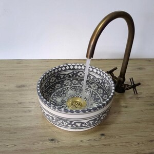 May include: A patterned ceramic sink with a brass tap. The sink has a blue and white design and is filled with water. The tap is vintage-style and water is flowing into the sink. The sink is on a wooden surface.