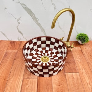May include: A round ceramic vessel sink featuring a brown and white checkered pattern on the interior and exterior. The sink includes a gold-toned metal drain and is paired with a tall, curved, antique-style brass faucet with two cross-handle knobs.