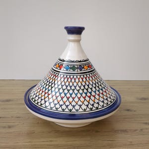 Puede incluir: Una olla de tagine de cerámica blanca con tapa cónica y base ancha. La tapa está decorada con un colorido patrón geométrico y la base tiene un borde azul. La olla está diseñada para cocinar y servir platos marroquíes.