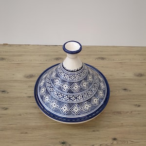 May include: A ceramic tagine pot with a conical lid and a circular base. The pot is white with intricate blue geometric patterns and a blue rim. The lid has a small, cylindrical handle. The tagine is displayed on a wooden surface.