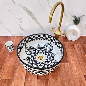 Puede incluir: Un lavabo de cerámica redondo con un patrón geométrico de mandala en blanco y negro, acompañado de un grifo alto de tono dorado y un pequeño vaso a juego. El lavabo cuenta con una válvula de desagüe dorada y un borde triangular.
