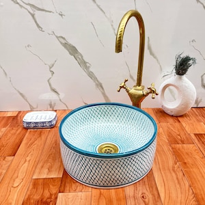 Könnte beinhalten: Ein rundes Keramik-Aufsatzwaschbecken mit einem blaugrün-weißen Gittermuster, kombiniert mit einer hohen Messingarmatur. Eine kleine rechteckige Seifenschale mit blau-weißem Muster und eine weiße, strukturierte runde Vase stehen daneben.