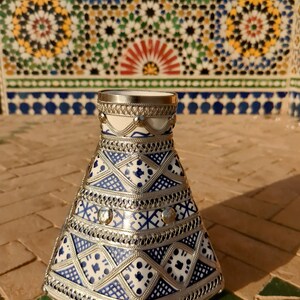 Puede incluir: Un jarrón decorativo de cerámica con base blanca y complejos diseños azules. El jarrón está adornado con filigranas y detalles de metal plateado. El diseño presenta formas geométricas y es adecuado para la decoración del hogar.