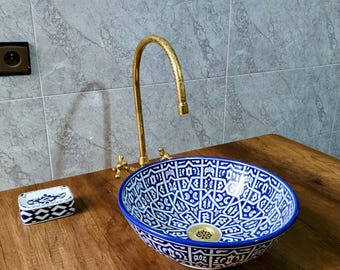 Può includere: Un lavabo decorativo in ceramica con motivi blu e bianchi con un rubinetto dorato. A sinistra c'è un portasapone abbinato. Il lavabo è rotondo e posto su una superficie di legno. Lo sfondo presenta piastrelle grigie.