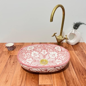 May include: A round ceramic vessel sink featuring a white base with intricate pink hand-painted floral and geometric patterns. The sink includes a gold-toned metal drain and is paired with a tall, curved, antique-style brass faucet.