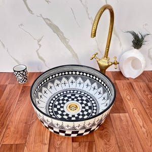 May include: A round ceramic vessel sink featuring a black and white geometric pattern with a central gold drain. The sink is paired with a tall, curved, antique-style brass faucet and a small matching black and white patterned ceramic cup.