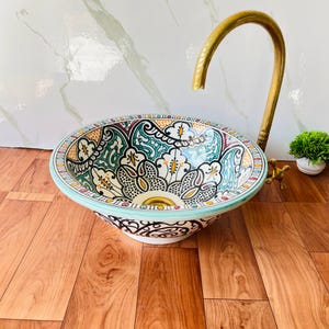 Puede incluir: Un lavabo de sobreponer redondo de cerámica con un intrincado patrón geométrico multicolor en tonos turquesa, amarillo, negro y rojo. Se combina con un grifo alto y curvo de latón cepillado con mandos en cruz.