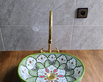 May include: Oval ceramic sink with a floral design in white, green, red, and black. The sink has a green border and is paired with a gold faucet. The sink is on a wooden surface.