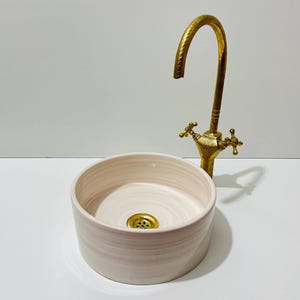 May include: A round, light pink ceramic sink with a gold-colored drain and a matching gold faucet. The sink has a simple, modern design, and the faucet features cross-shaped handles.