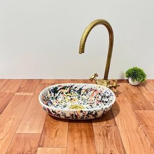 Puede incluir: Un lavabo ovalado de cerámica con base blanca y un patrón de salpicaduras de pintura multicolor en azul, rosa, amarillo y negro. El lavabo incluye un desagüe de metal dorado y se combina con un grifo alto y curvo de latón antiguo.