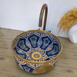 Peut inclure: Un évier rond en céramique décoratif avec un motif floral bleu, blanc, orange et vert. L'évier a un bord et un couvercle de drain dorés. Un robinet en bronze est fixé à l'évier.