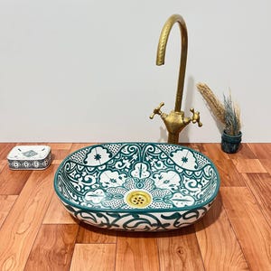 May include: An oval ceramic vessel sink featuring a white base with intricate dark green floral and geometric patterns. The sink includes a gold-toned drain, a tall brass faucet with two handles, a small matching soap dish, and a tiny decorative pot.