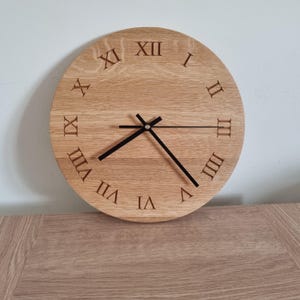 May include: A circular wall clock made of light brown wood with a visible grain pattern. The clock face features engraved Roman numerals from I to XII arranged around the perimeter. Three thin black metal hands are centred on the clock face.