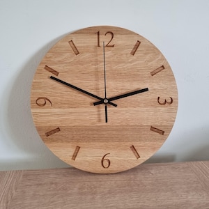 Op de afbeelding: Een ronde houten wandklok met een lichtbruine afwerking. De wijzerplaat heeft gegraveerde uurmarkeringen en zwarte wijzers. De cijfers 12, 3, 6 en 9 zijn zichtbaar. De klok is aan een witte muur bevestigd.