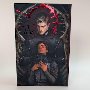 May include: A book cover featuring two figures in dark attire. One figure stands behind the other, who is resting with closed eyes. The artwork has a dark, gothic aesthetic with red accents and intricate details.