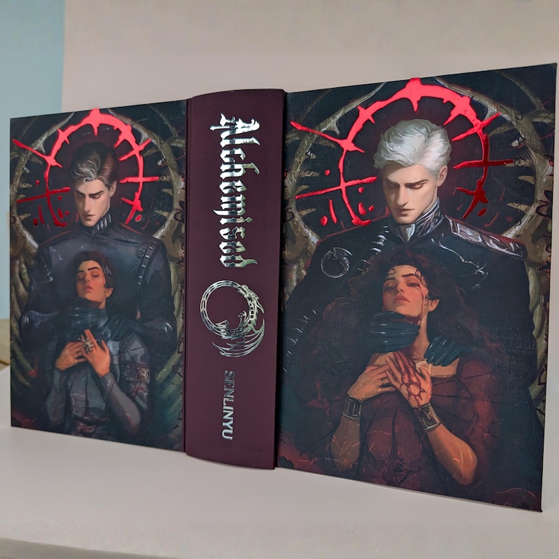 May include: A hardcover book with a burgundy spine and cover art featuring two figures. The title "Alchemist" is displayed vertically in silver lettering. The cover art depicts a man and woman in a dramatic pose, with a red, circular design.