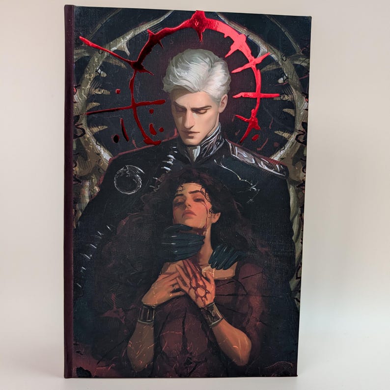 May include: A hardcover book with a dark, gothic-style illustration. The cover features a man with white hair and a woman, both with pale skin. Red accents and a dark background create a dramatic effect. The book's spine is a deep burgundy color.