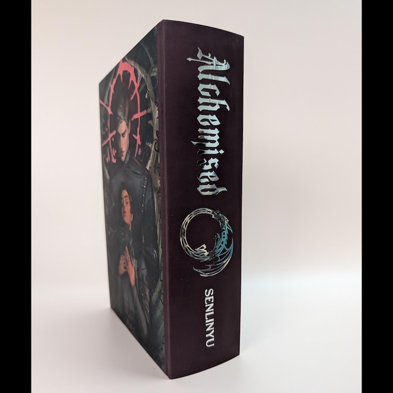May include: A hardcover book with a dark burgundy cover. The spine features the title "Alchemised" in silver lettering, along with the author's name, "SENLINYU." The book's cover art depicts two figures in dark clothing.
